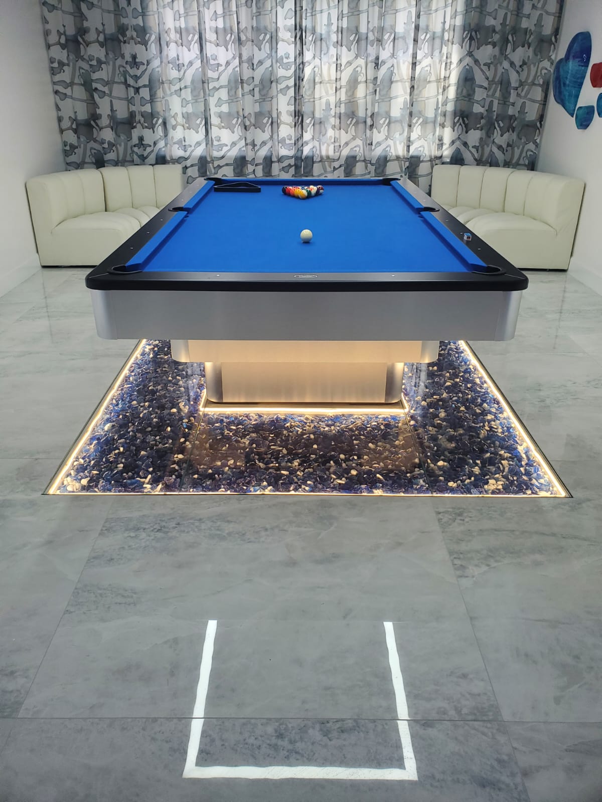A modern pool table with a blue felt top stands over a lit, glass-covered display of blue stones.