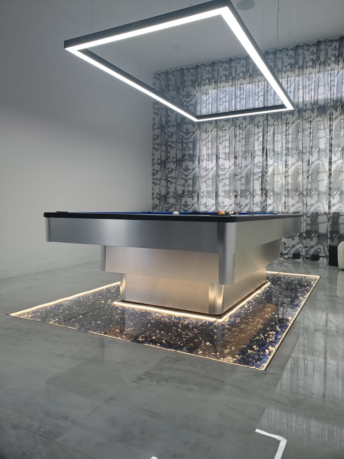 Modern pool table with a metallic finish, LED lighting, and glass stones underneath, in a sleek, bright room.