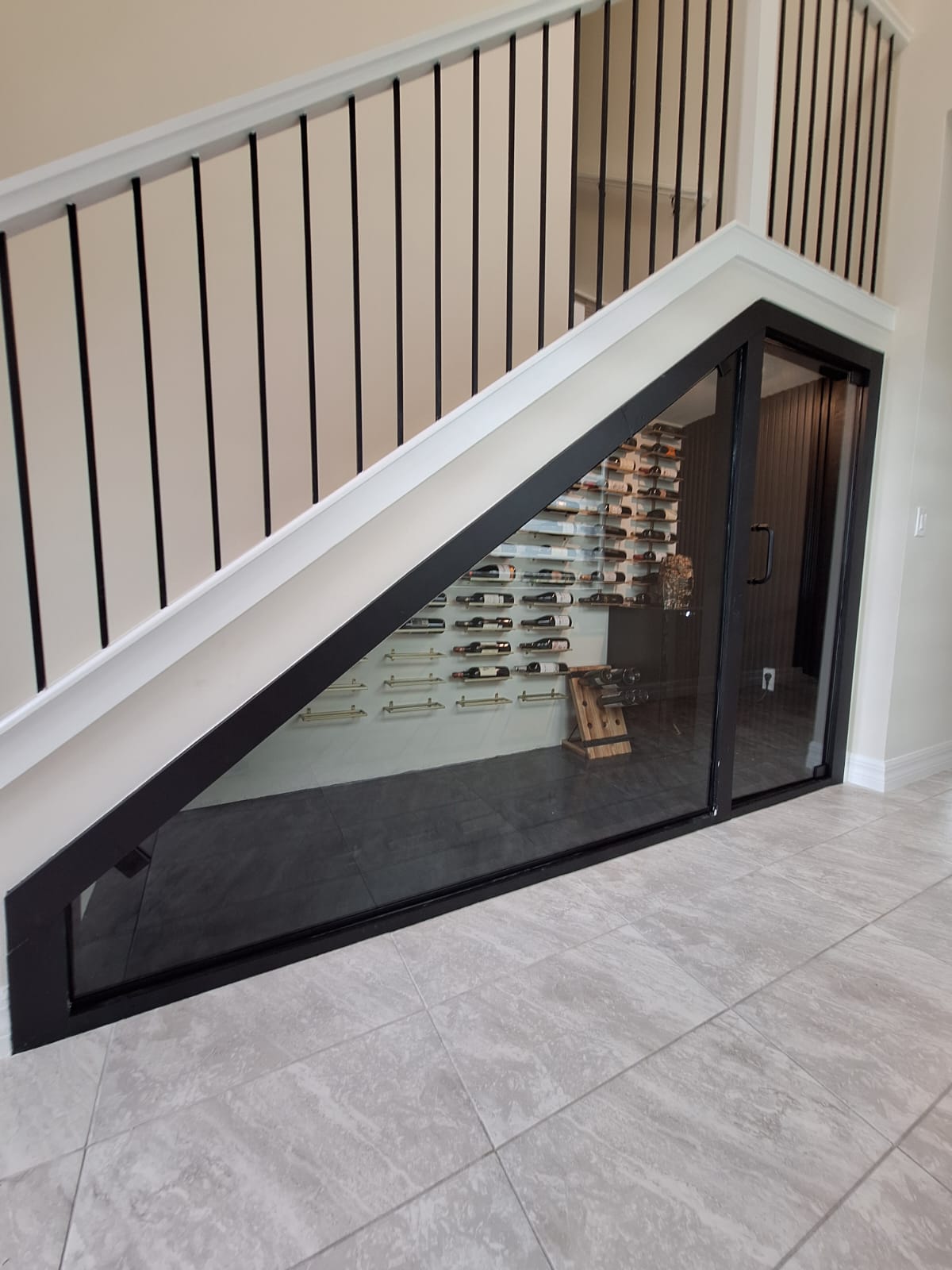 Modern under-stair wine cellar with glass doors, wine bottles on wall racks, and a tiled floor.