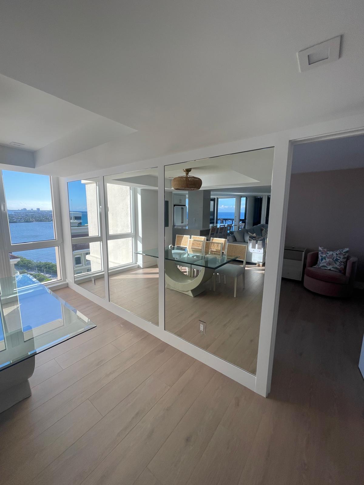 Bright dining area with large windows, glass table, and mirrored wall reflecting natural light and outdoor view.