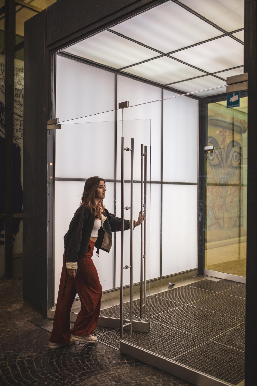 A woman in red pants opens a glass door to a brightly lit, modern building entrance.