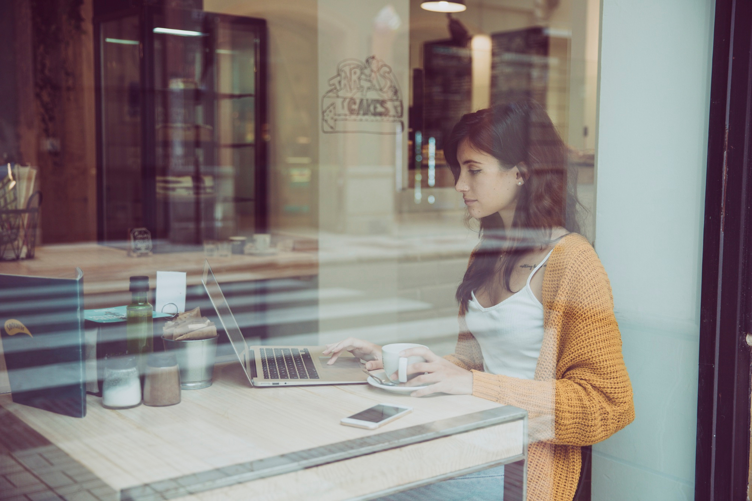 Woman sitting in a café, using a laptop and holding a cup, seen through a window.